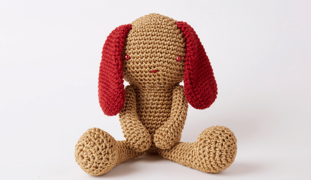 A photo of a crochet plushie bunny with red ears and a beige body.