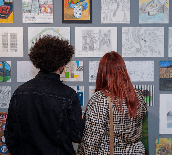 A photo of a couple standing in front of a wall of artwork at a Koestler Arts exhibition.