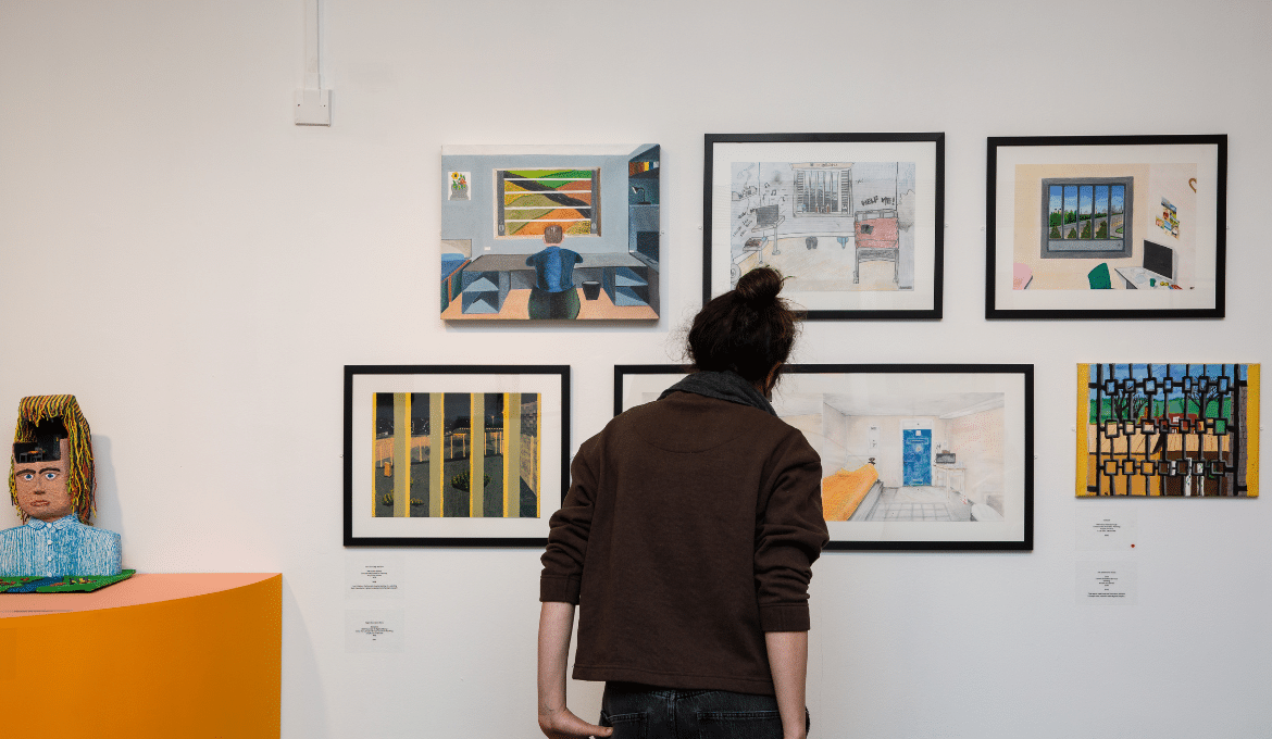 A photo of a person stood in front of a wall full of artwork at a Koestler Arts exhibiton.