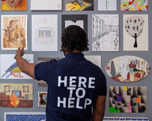A Koestler Arts volunteer stood in front of an exhibition wall with a 'HERE TO HELP' shirt on.