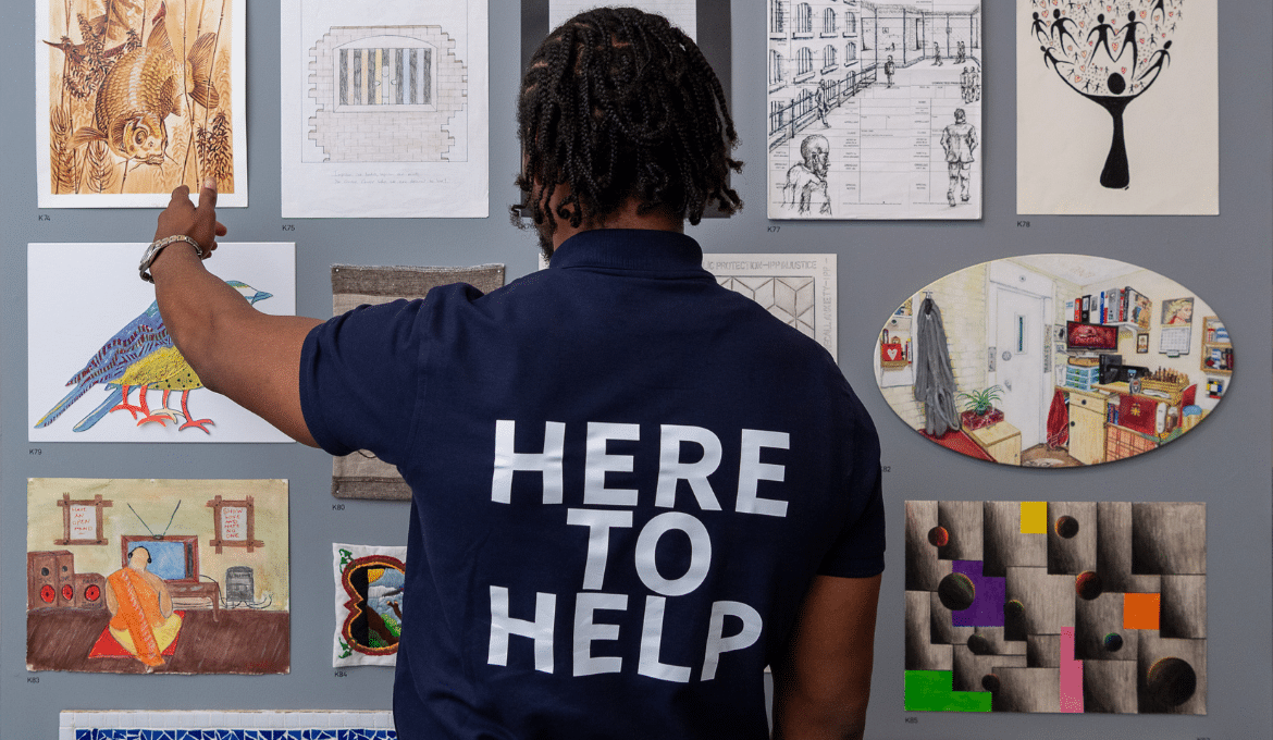 A photo of the back of a Koestler Arts volunteer in front of a wall of artwork with a top that says 'Here to help'.