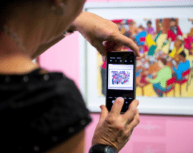 A photo of someone taking a picture on their phone of an artwork at a Koestler Arts exhibition.